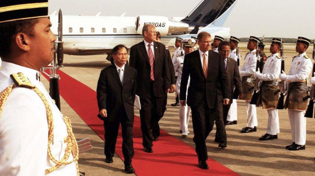 A picture of Andrew Mountbatten-Windsor arriving in Kuala Lumpur flanked by Malaysian soldiers and walking along a red carpet in 2007. He is wearing a dark shit and pink tie, accompanied by two other men also wearing suits.