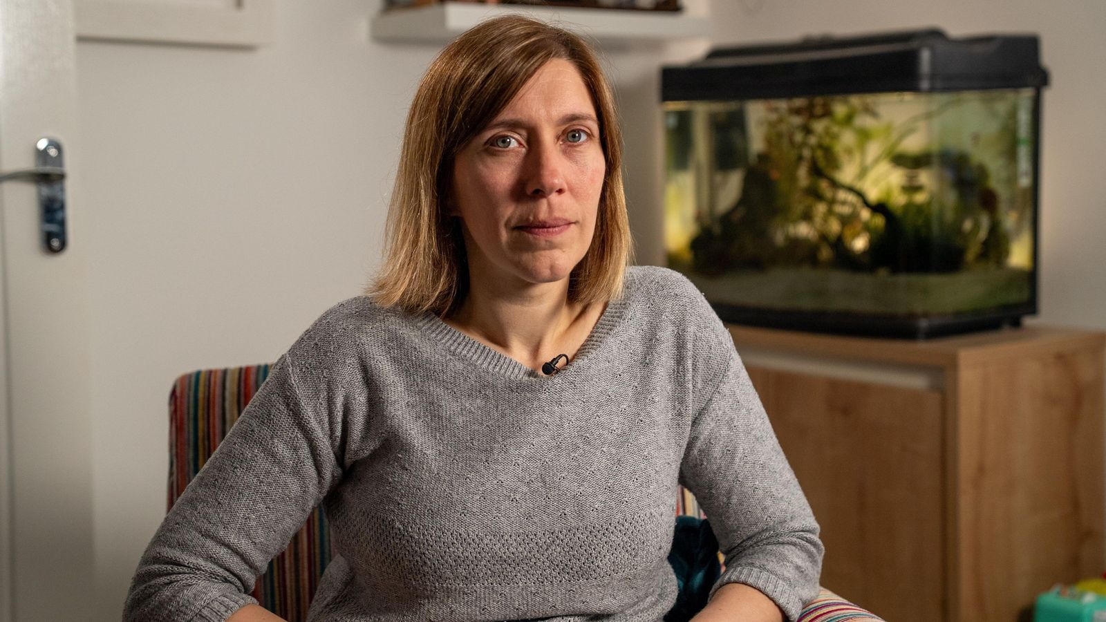 Catriona Ball sits on a striped armchair. There is a brown cupboard and fish tank behind her. She is wearing a grey jumper. She has brown/blonde hair in a bob.