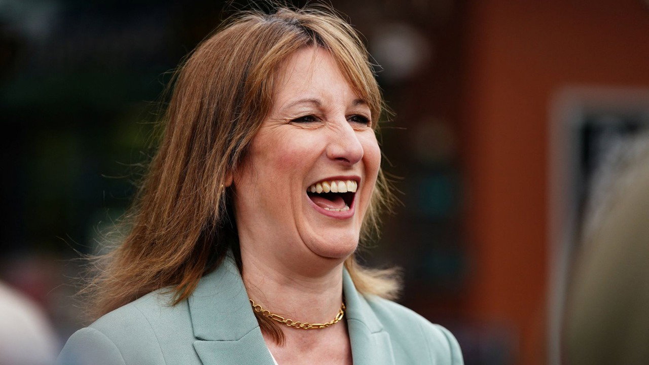 Rachel Reeves laughs at something off camera. She is wearing a teal-green suit jacket and a gold chain necklace.