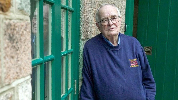 An older man with grey receding hair and glasses leans against a brick wall looking towards the camera. He wears a blue sweatshirt. He looks sad.
