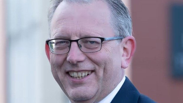 A man with short grey hair and glasses and wearing a dark blue suit, white shirt and light blue tie is smiling.