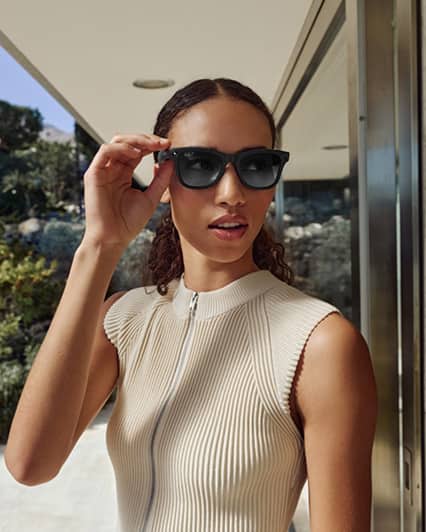 Person with long hair wearing sunglasses and a sleeveless, ribbed top, standing outdoors near a glass building. They hold the sunglasses with one hand, with greenery visible in the background.