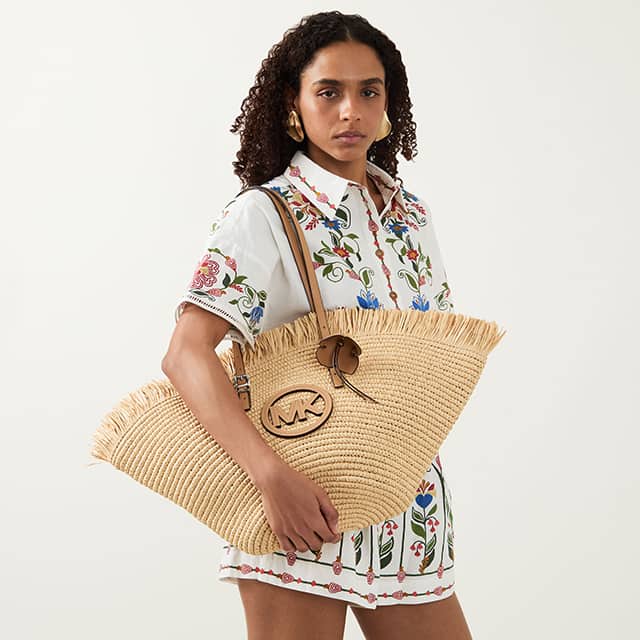 A woman with curly hair wearing a white floral-patterned shirt dress holds a large woven straw handbag with a circular logo and tassel. She stands against a plain background, showcasing her outfit and accessories, including large hoop earrings.