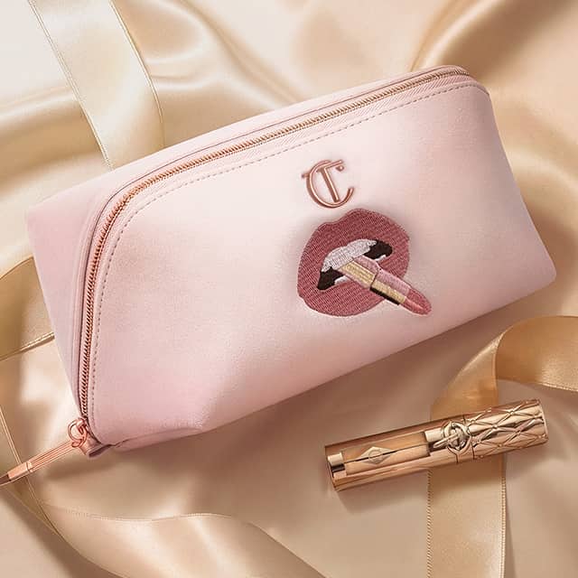 A pink makeup pouch featuring an embroidered design of lips applying lipstick, with a metallic zipper and a logo emblem. A gold lipstick tube lies nearby on a smooth satin fabric surface.