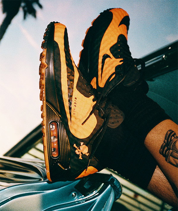 Orange and black Nike Air Max trainers with visible Air unit and skull logo, worn by person with leg tattoo resting on car door