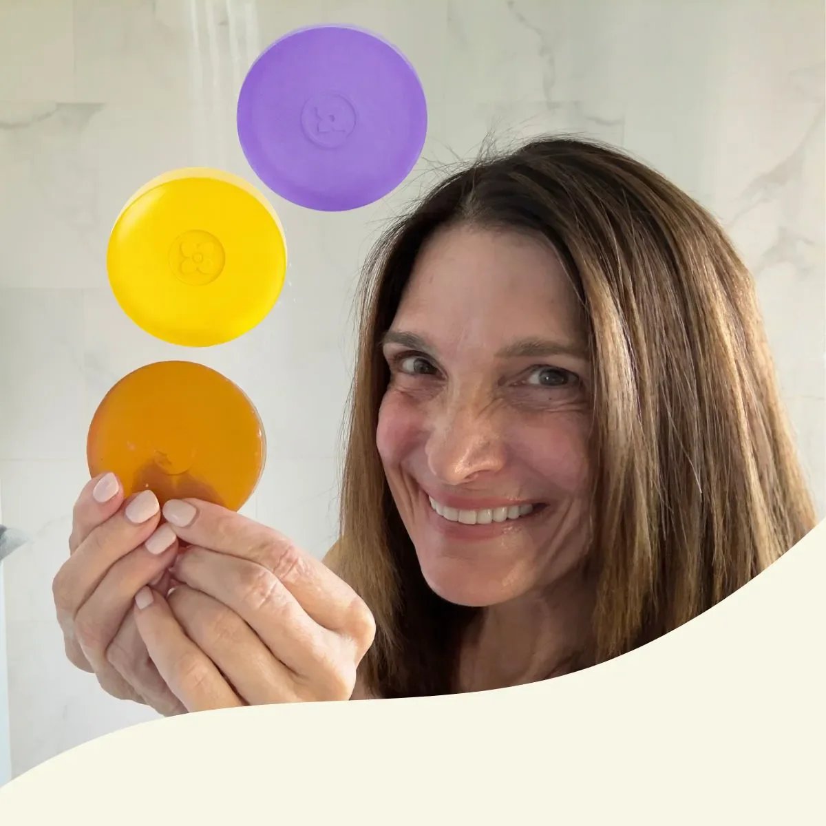 A smiling woman holds up three round bars of soap in yellow, orange, and purple colors. The background appears to be a shower with water falling. (square)