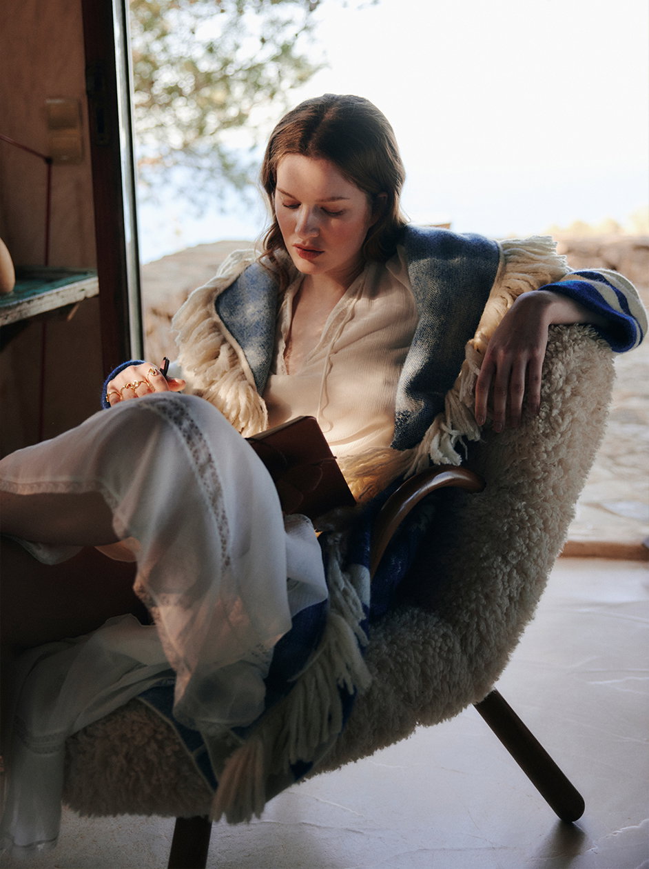 Eine Frau sitzt entspannt in einem flauschigen Sessel, trägt ein zartes weißes Spitzenkleid und einen blau-beige gestreiften Cardigan mit langen Fransen von DOROTHEE SCHUMACHER und schreibt in ein Notizbuch.