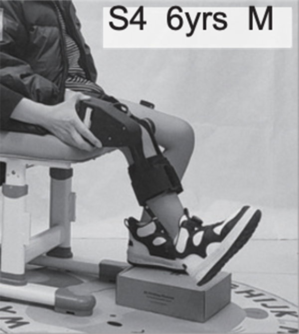 A child sitting on a chair extends one of his legs with the help of a robotic device worn around it.