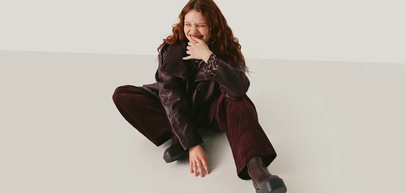 Joven mujer con cabello rizado rojo sentada en el suelo con pantalones y chaqueta de color púrpura oscuro, sonriendo y tocándose la cara sobre un fondo neutro.