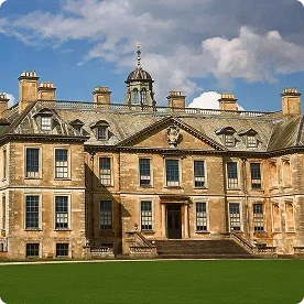 A large, historic stone manor house with a grand staircase and a manicured green lawn under a cloudy sky.