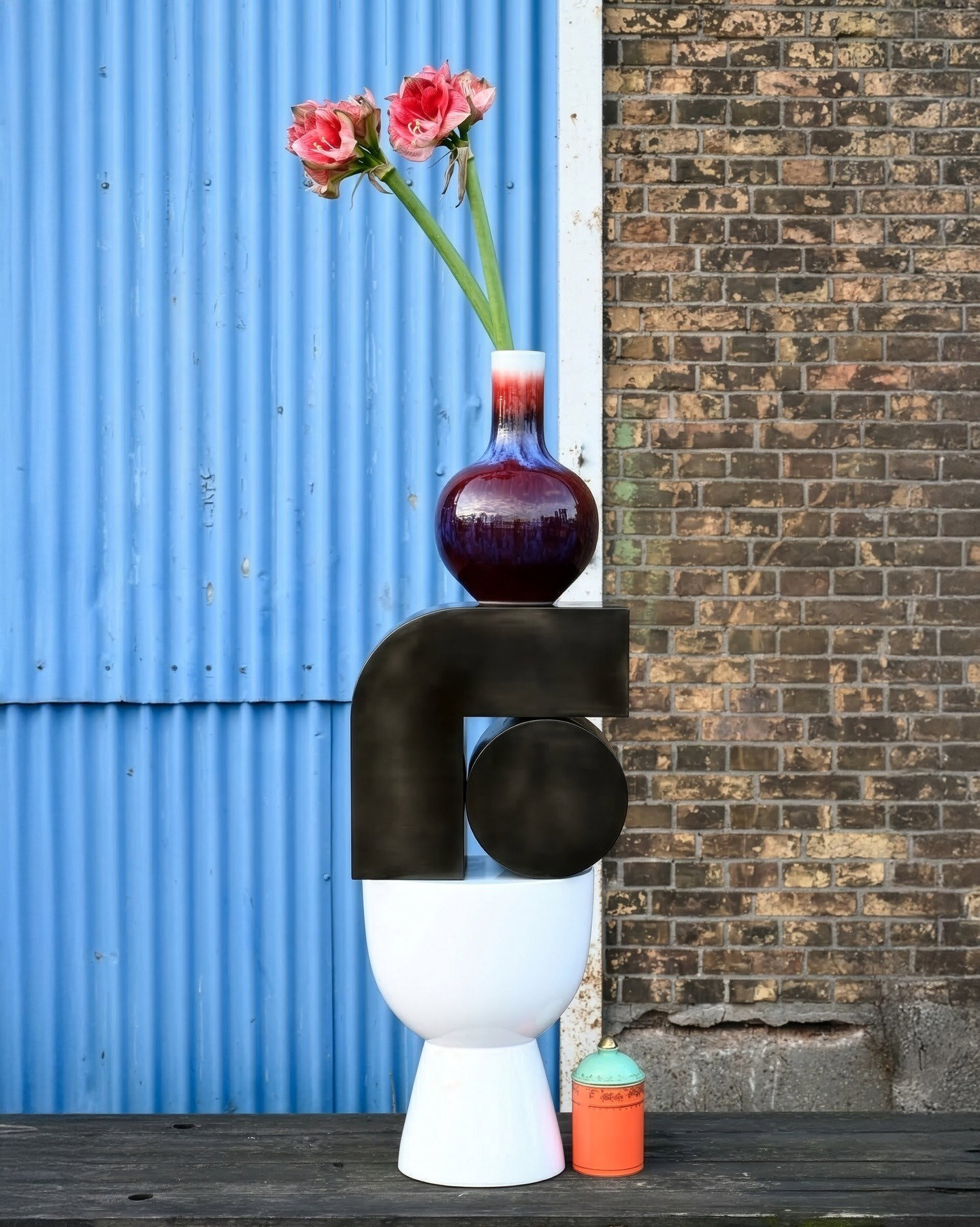 Modern still-life: red glass vase with pink amaryllis on stacked sculptures against blue corrugated metal and brick wall