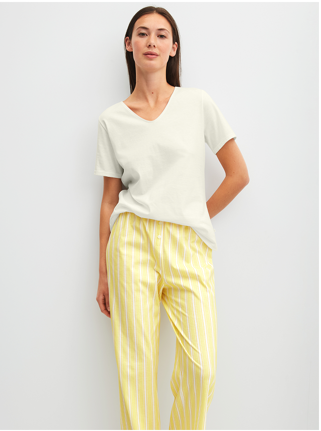 The model is wearing striped 7/8-length pajama pants in the color “Cream Gold” and a V-neck T-shirt in the color “Star Dust,” both from CALIDA's FAVOURITES OCEAN collection. 