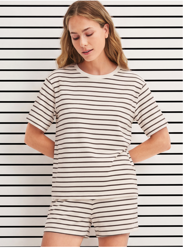 The model is wearing a striped short-sleeved shirt and striped shorts, both in the color “Charm Cream” and from the FAVOURITES LAYERING collection by CALIDA.