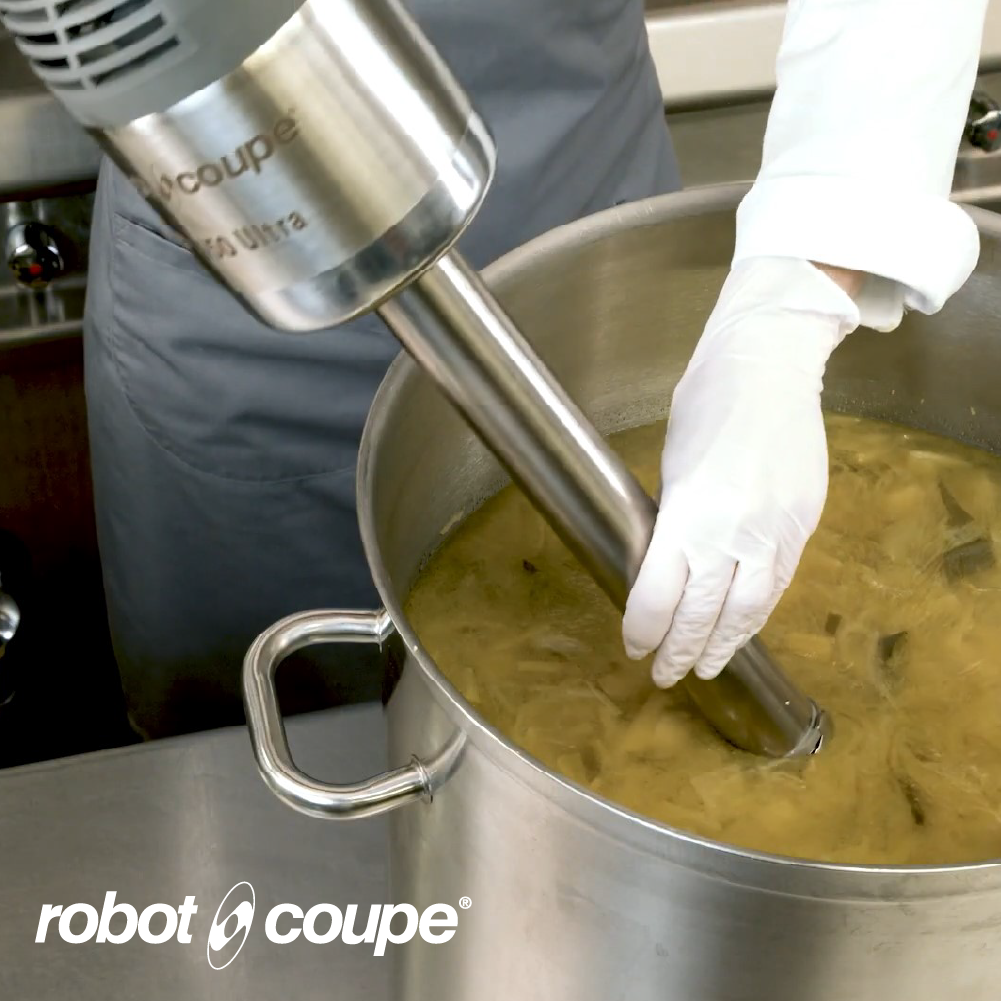 Robot Coupe immersion blender mixing soup inside a large stainless steel stock pot in a commercial kitchen.