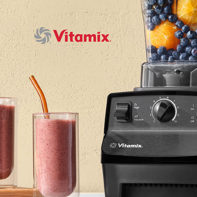 Vitamix commercial blender filled with fresh fruit ingredients beside two finished smoothies in clear glasses.