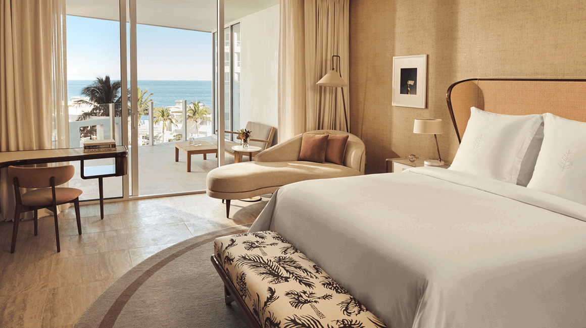 Ocean-view bedroom with a king bed, chaise lounge, and desk, opening onto a balcony with seating and views of palm trees and the sea.