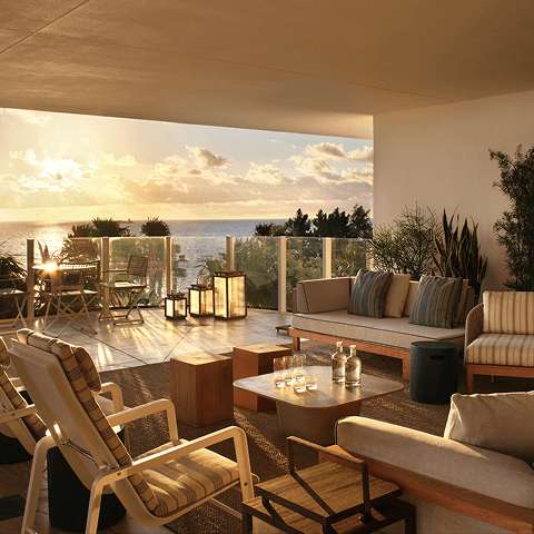 Hotel room interior opening onto a sunlit balcony with table and chairs overlooking the ocean.