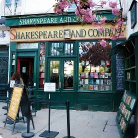 The green storefront of the Shakespeare and Company bookstore in Paris, framed by blooming pink flowers.
