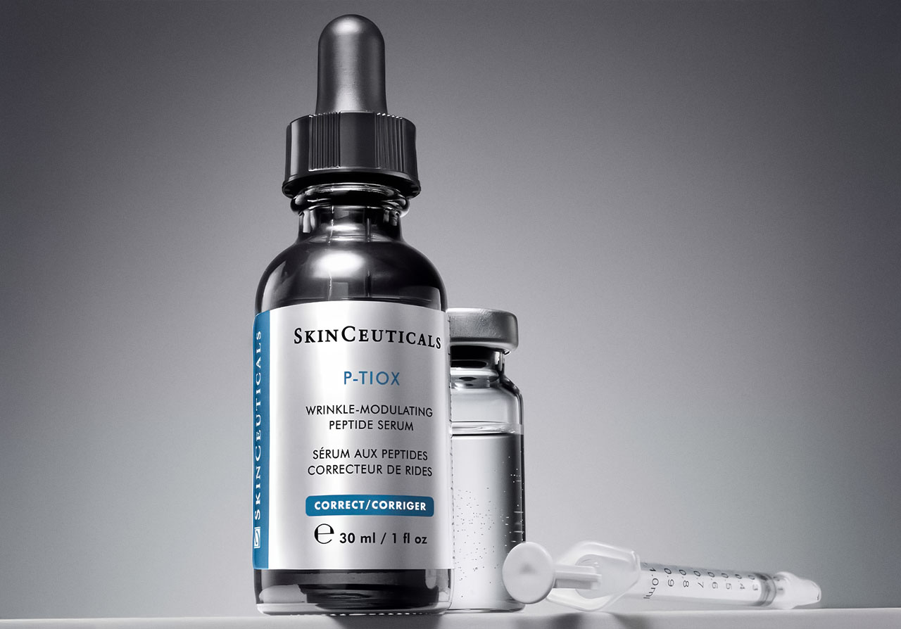 30ml Flasche des SkinCeuticals P-TIOX Serums. Direkt daneben sieht man eine kleine gläserne Ampulle mit klarer Flüssigkeit und eine medizinische Spritze (ohne Nadel), die flach auf dem Boden liegt.