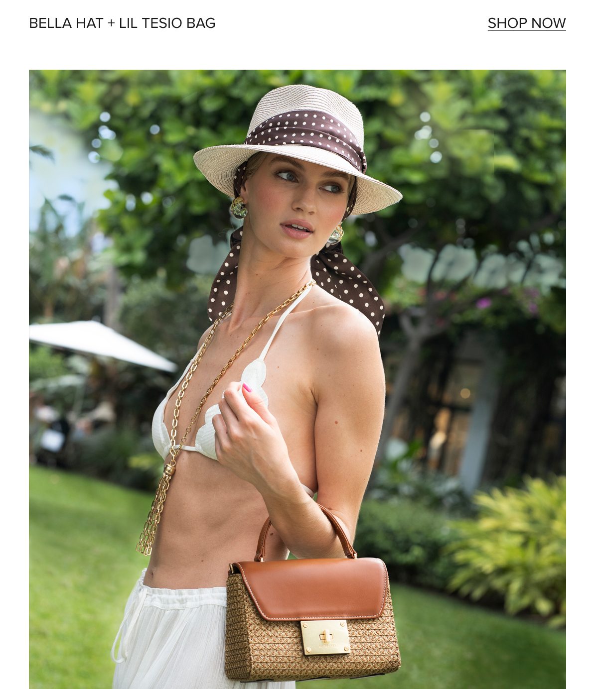 A woman in a bikini and hat holds a stylish handbag in a lush outdoor setting.