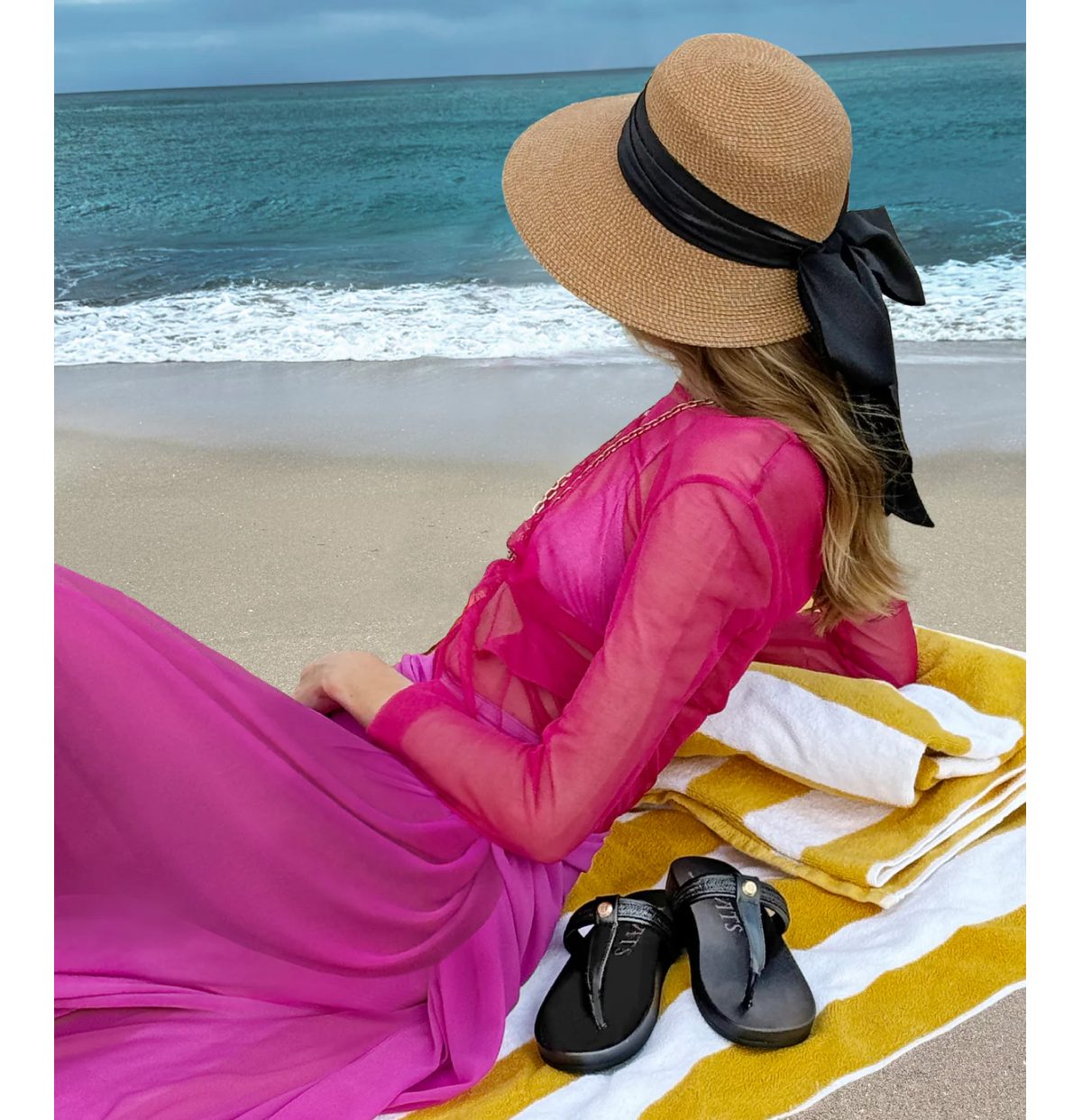 Woman in a pink dress and straw hat relaxing on a beach towel by the ocean.