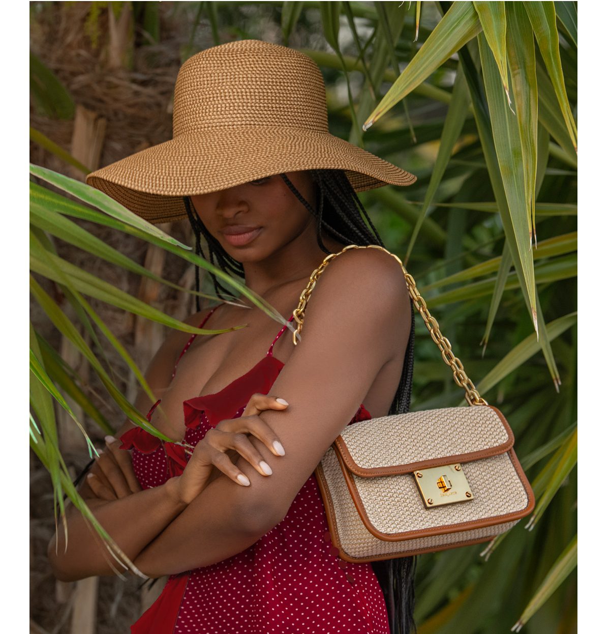 A woman in a wide-brimmed hat and red dress poses with a stylish handbag among green palm leaves.