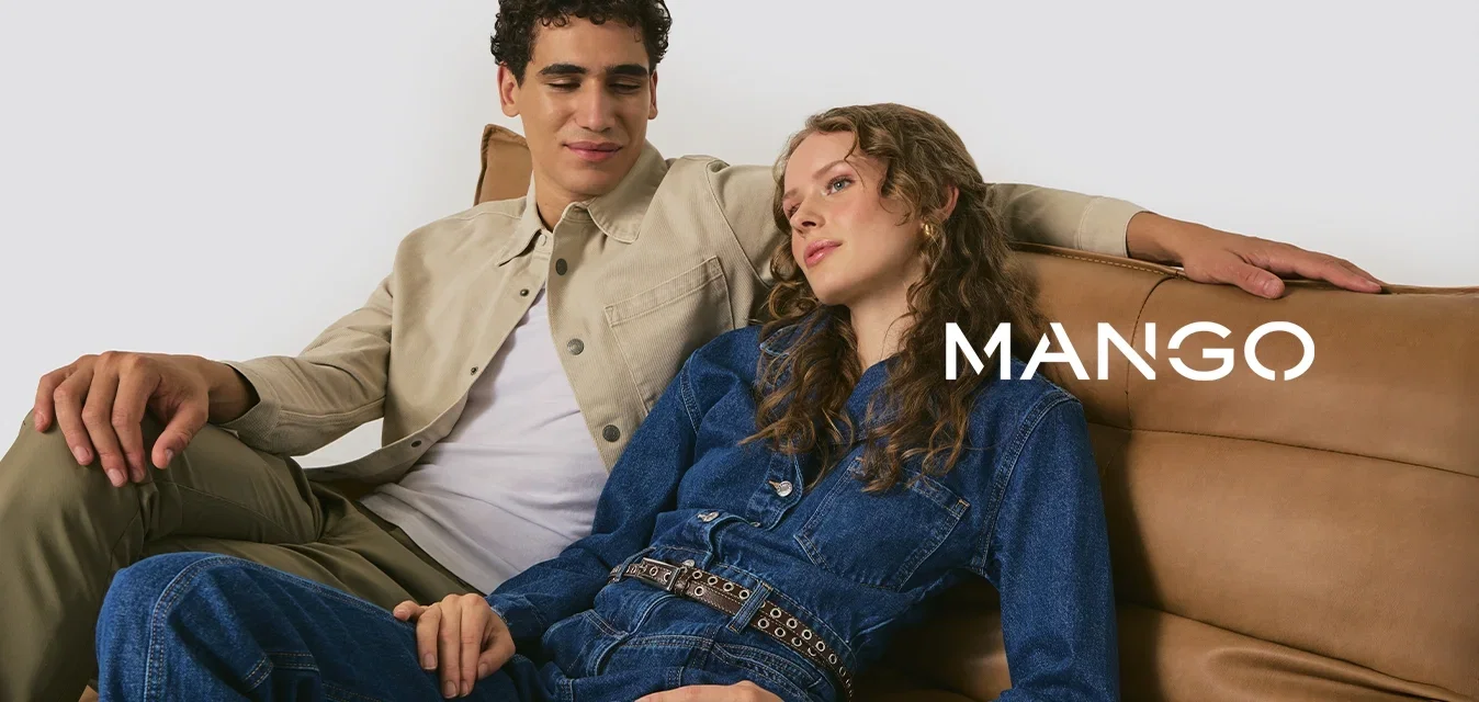 Twee personen zitten op een lichtbruin leren sofa. De ene draagt een beige jas over een wit shirt, de ander een blauwe denim jumpuit met een riem.
