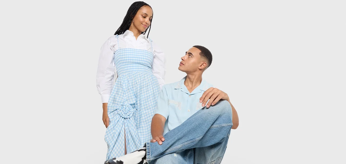 Vrouw in een blauw geruite jurk staat naast een man die op een rode stoel zit, gekleed in een lichtblauw shirt en jeans, terwijl ze naar elkaar kijken.