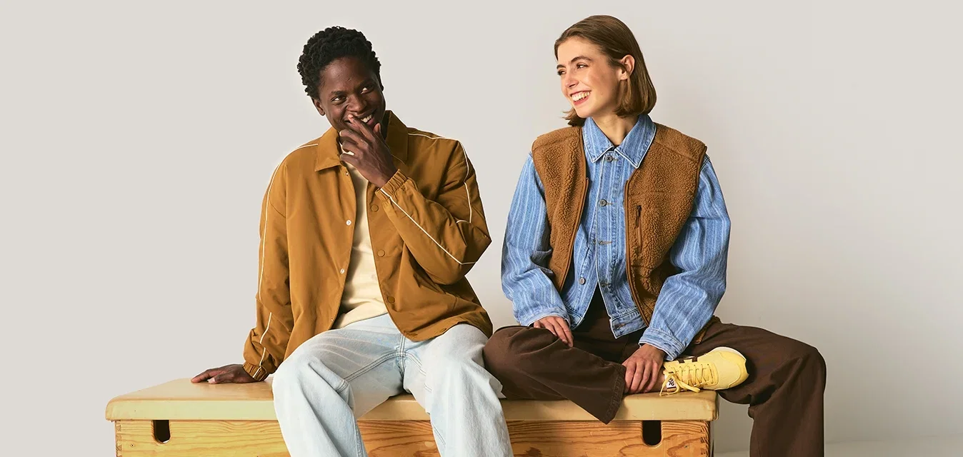 Dos personas sentadas en un banco de madera. Una lleva una chaqueta marrón sobre una camisa beige; la otra luce una camisa azul y un chaleco marrón. Son visibles unas zapatillas deportivas.