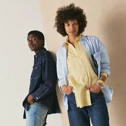 Deux personnes sont appuyées de manière décontractée contre un mur ; l'une porte une chemise en jean bleue et un jean, l'autre une chemise bleu clair, un haut jaune et un jean.