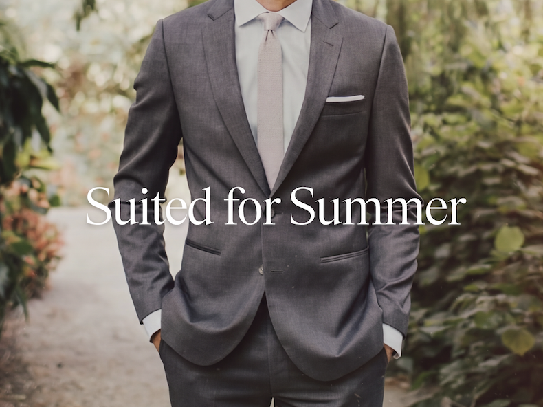 Man wearing a charcoal gray suit with a light gray dress shirt, tan tie, and white pocket square, standing outdoors among lush green foliage. Suited for Summer