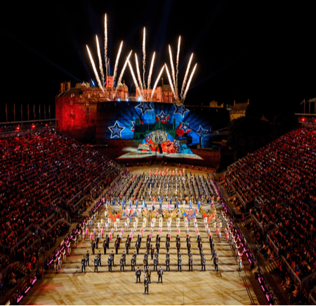 A vibrant night scene of a large outdoor performance with fireworks and a historic castle backdrop.