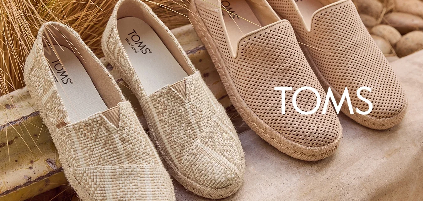 Twee paar beige instapschoenen, één met een geweven textuur en de ander met perforaties, geplaatst op een stenen oppervlak buiten, met droge grassen erachter.