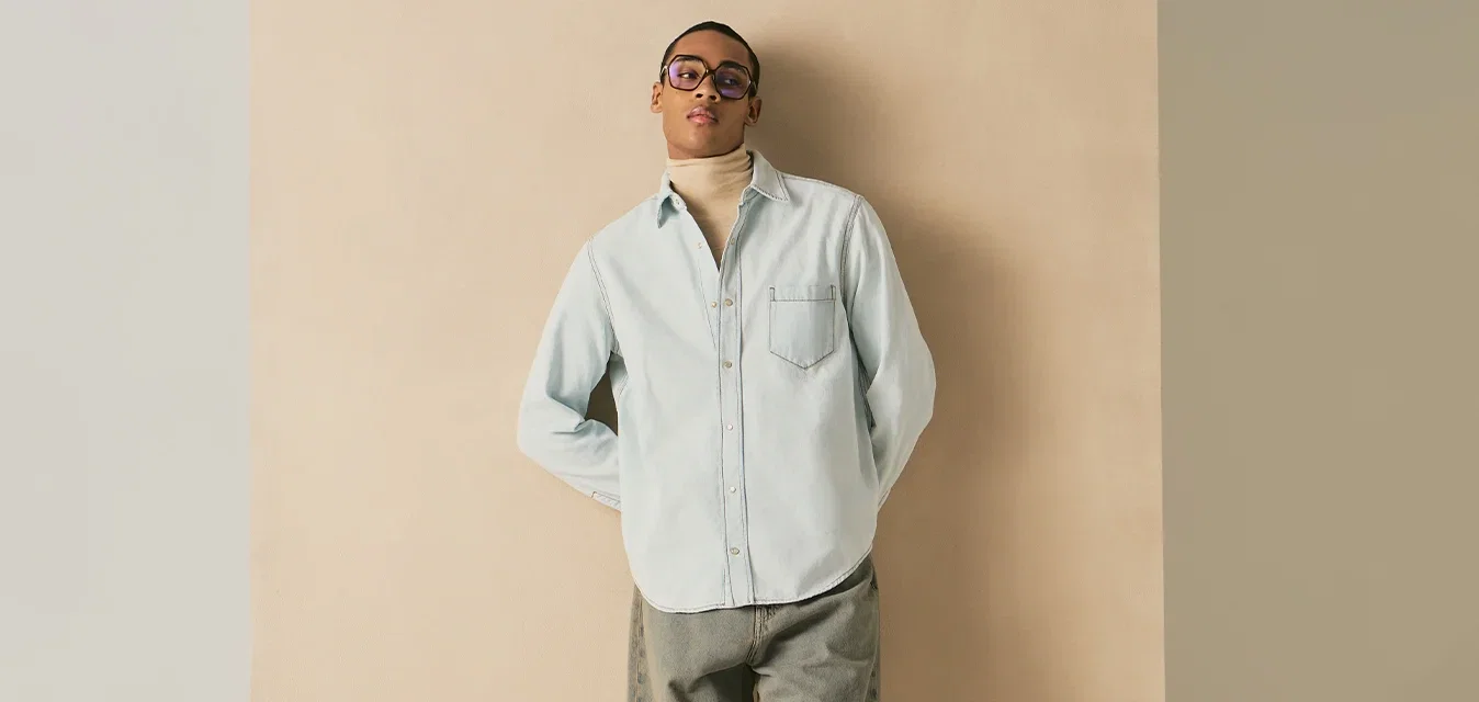 Jonge man met bril, licht denim overhemd over een beige coltrui, losse kaki broek, staand tegen een beige achtergrond met handen achter de rug.