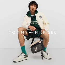 Model zit op een stoel, draagt een crèmekleurige bomberjack, een groene hoodie, zwarte shorts en witte sneakers, en houdt een kleine zwarte tas vast.
