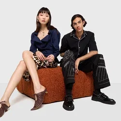 Twee personen zitten op een getextureerd bruin zitmeubel. De ene draagt een blauwe shirt en een patroonbroek; de ander een zwart shirt en gestreepte broek.