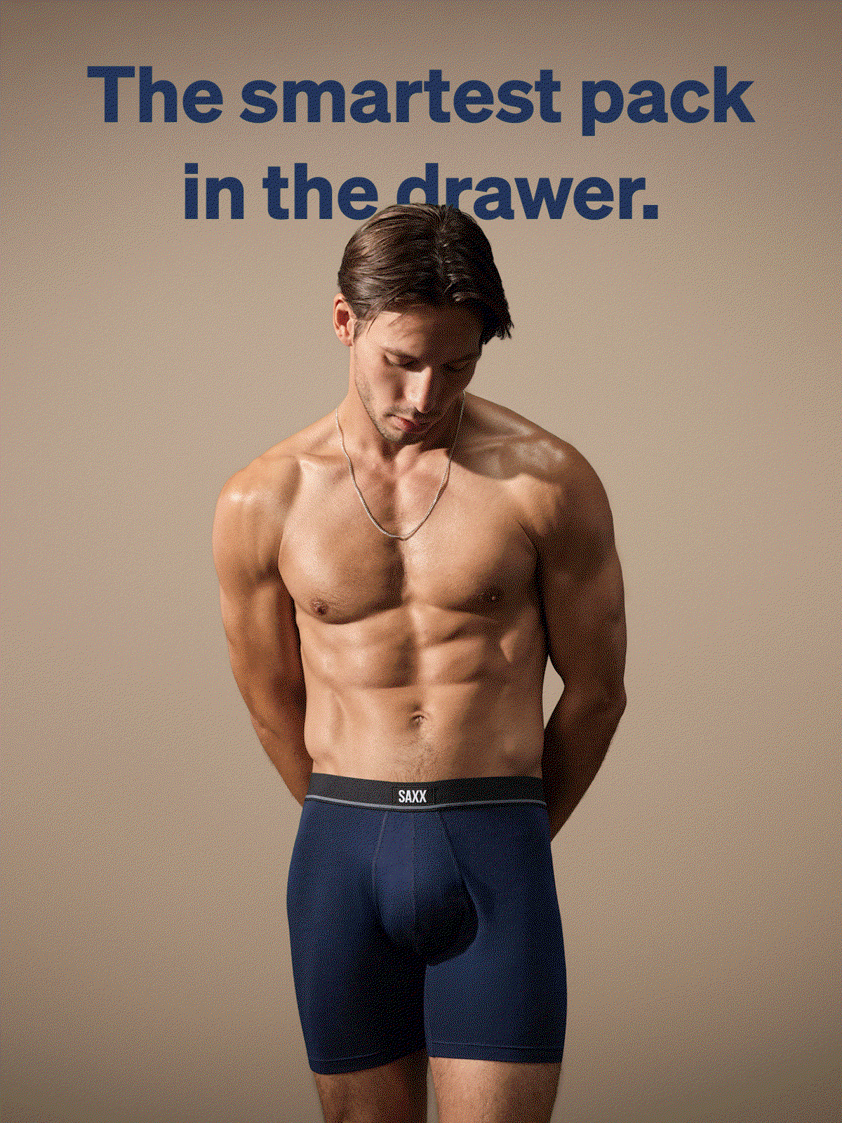 An athletic man wearing a silver chain and navy blue SAXX boxer briefs, looking down with hands behind his back.