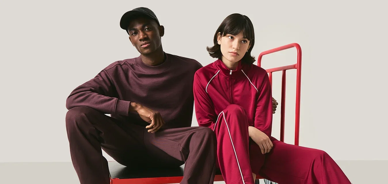 Zwei Personen in lässigen, passenden burgunderroten Outfits sitzen auf und neben einem roten Plattform-Handwagen vor einem neutralen Hintergrund.