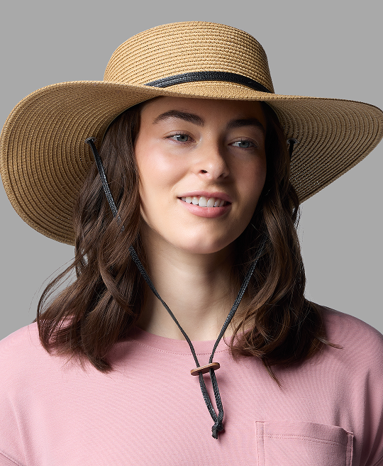 A woman wearing a wide-brimmed straw hat and a pink shirt against a grey background.