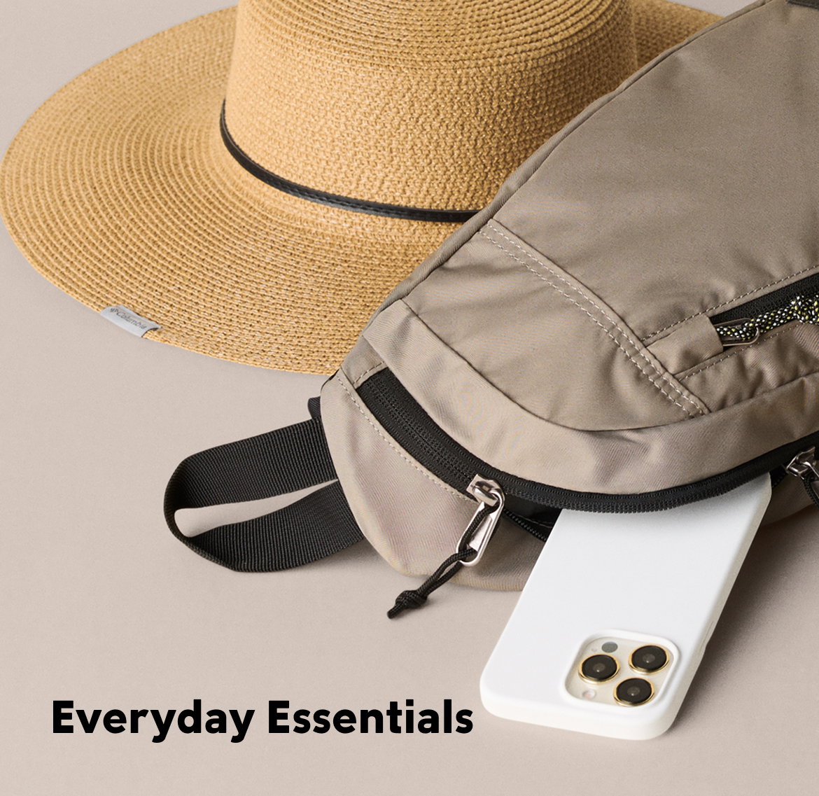 Close-up of a tan cross body bag on its side with a white iPhone slipping out, as well as a straw sun hat next to it, all against a tan backdrop with text: Everyday Essentials