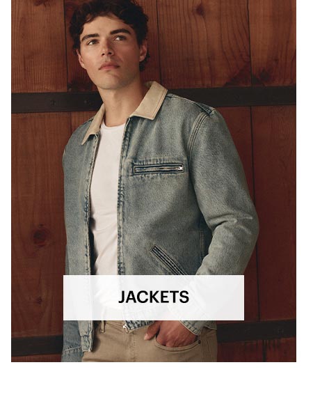 Man wearing a light wash denim zip-up jacket with a beige collar over a white tee and tan jeans, leaned against a dark wood paneled door. Shop men's jackets.