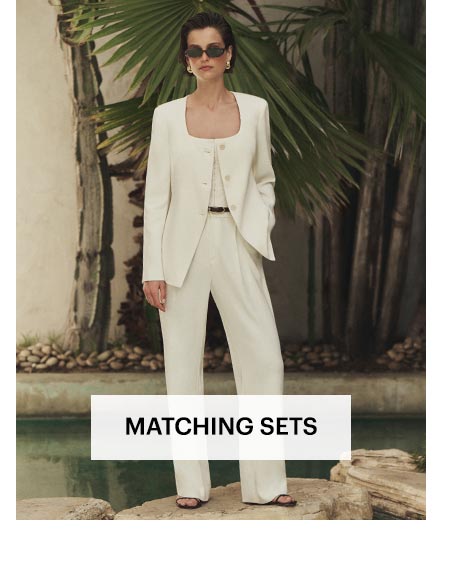 Woman wearing a white scoop neck blazer and trouser set, stood on a large stepping stone in a sunlit area surrounded by water and greenery. Shop women's matching sets.