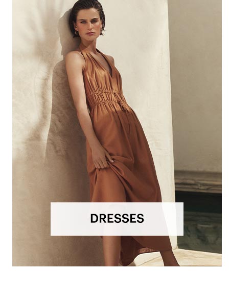 Woman wearing a burnt-orange halter midi dress, leaned against a stone wall in a sunlit area. Shop women's dresses.