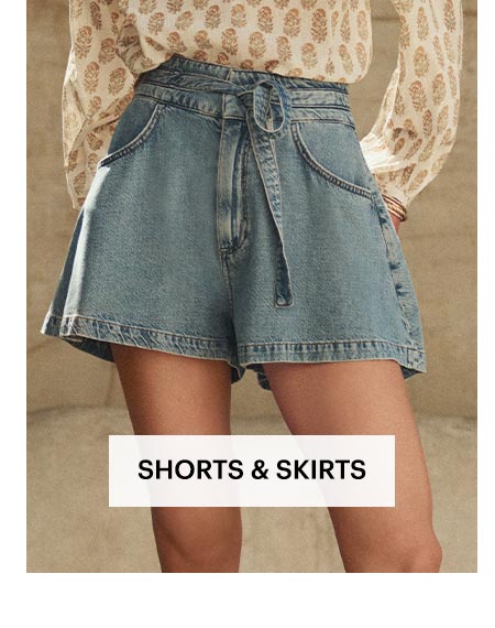 Woman wearing a pair of drapey, medium wash tie-waist denim shorts in the sunny outdoors. Shop women's shorts and skirts.