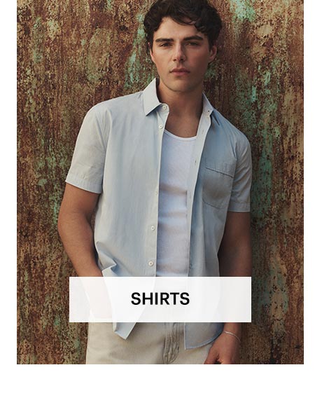 Man wearing a light bluish grey short sleeve button up over a white tank with a pair of off-white jeans, standing against an earthy textured wall. Shop men's shirts.