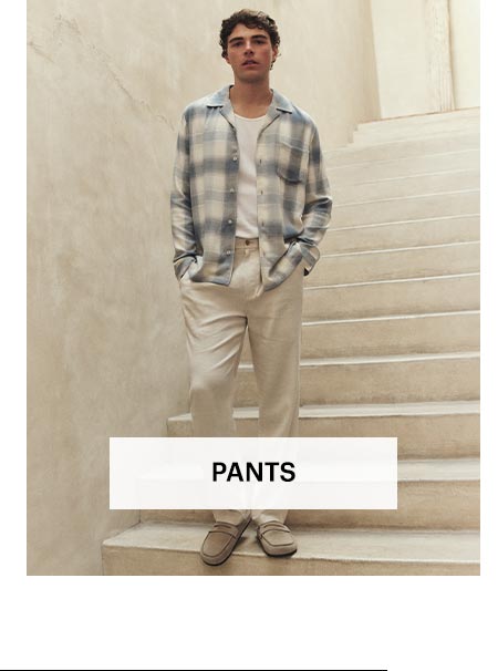 Man wearing cream colored pants with a white tee under a blue and white plaid long sleeve button up, stood on a sunlit stone staircase. Shop men's pants.