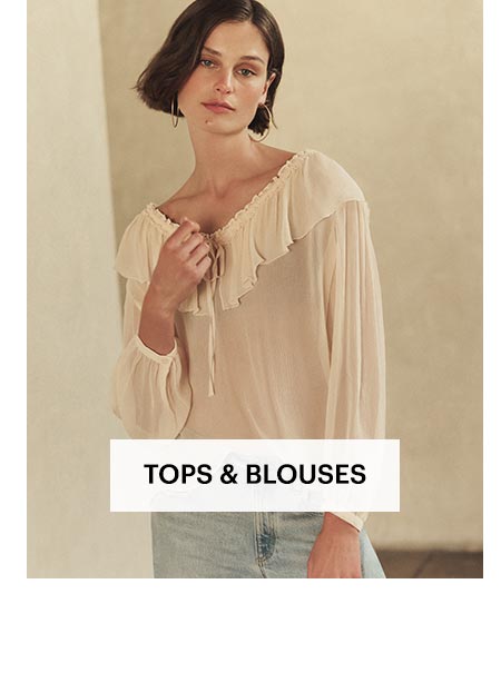 Woman in a light peach long sleeve silk blouse with light wash high rise jeans, posed in front of a sunlit stone wall. Shop women's tops and blouses.