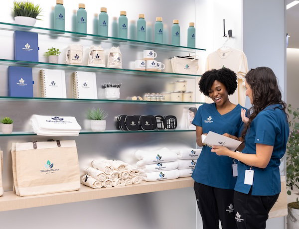 Two women wearing Blissful Roots branded clothing standing in front of shelves filled with Blissful Roots branded merch.