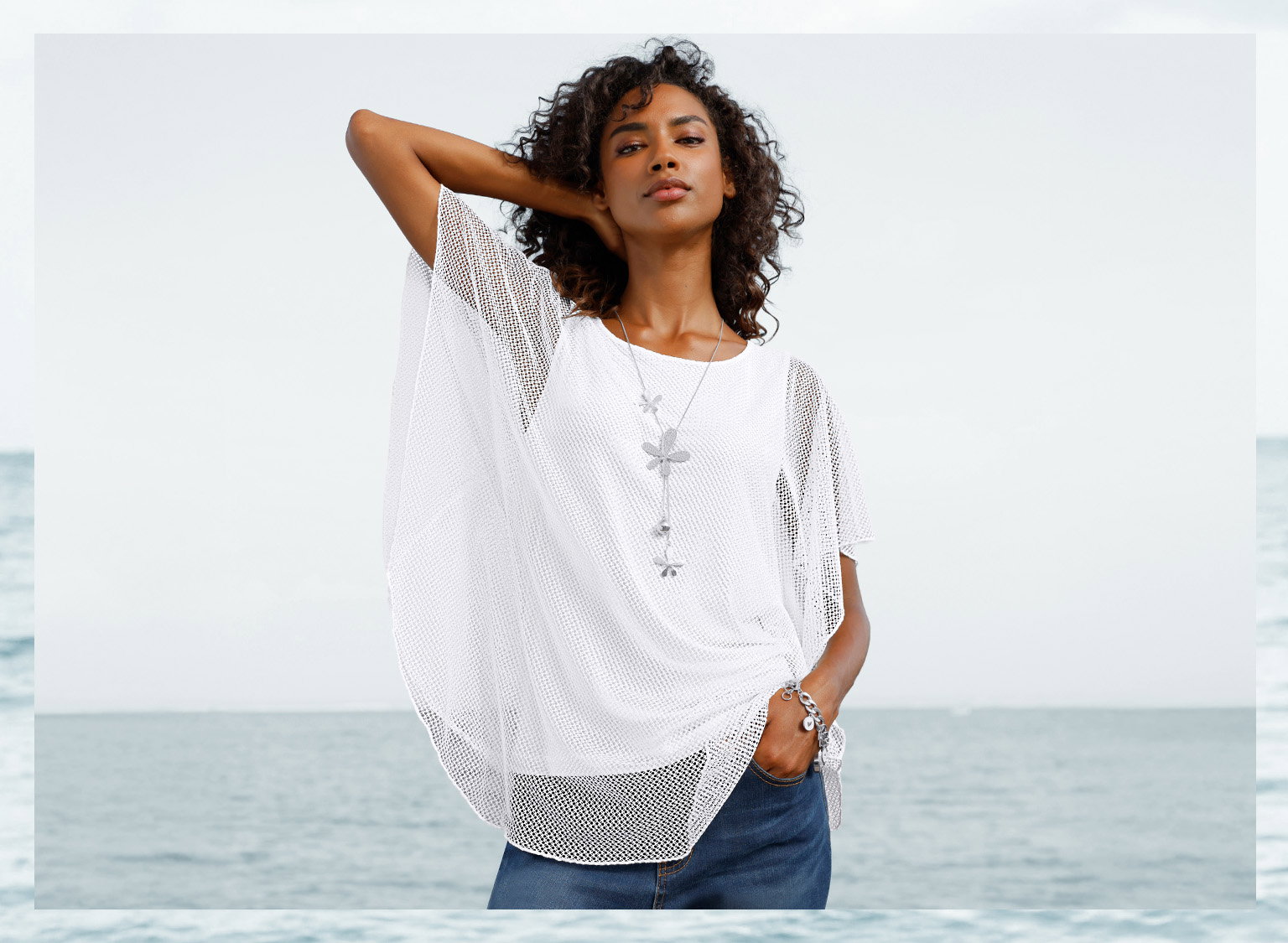 Een model draagt een wit, losvallend nettop met korte mouwen en sieraden, met de zee op de achtergrond.