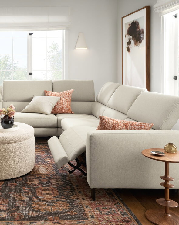 Elio sectional with cream upholstery and reclining footrest beside a round ottoman and wood table.